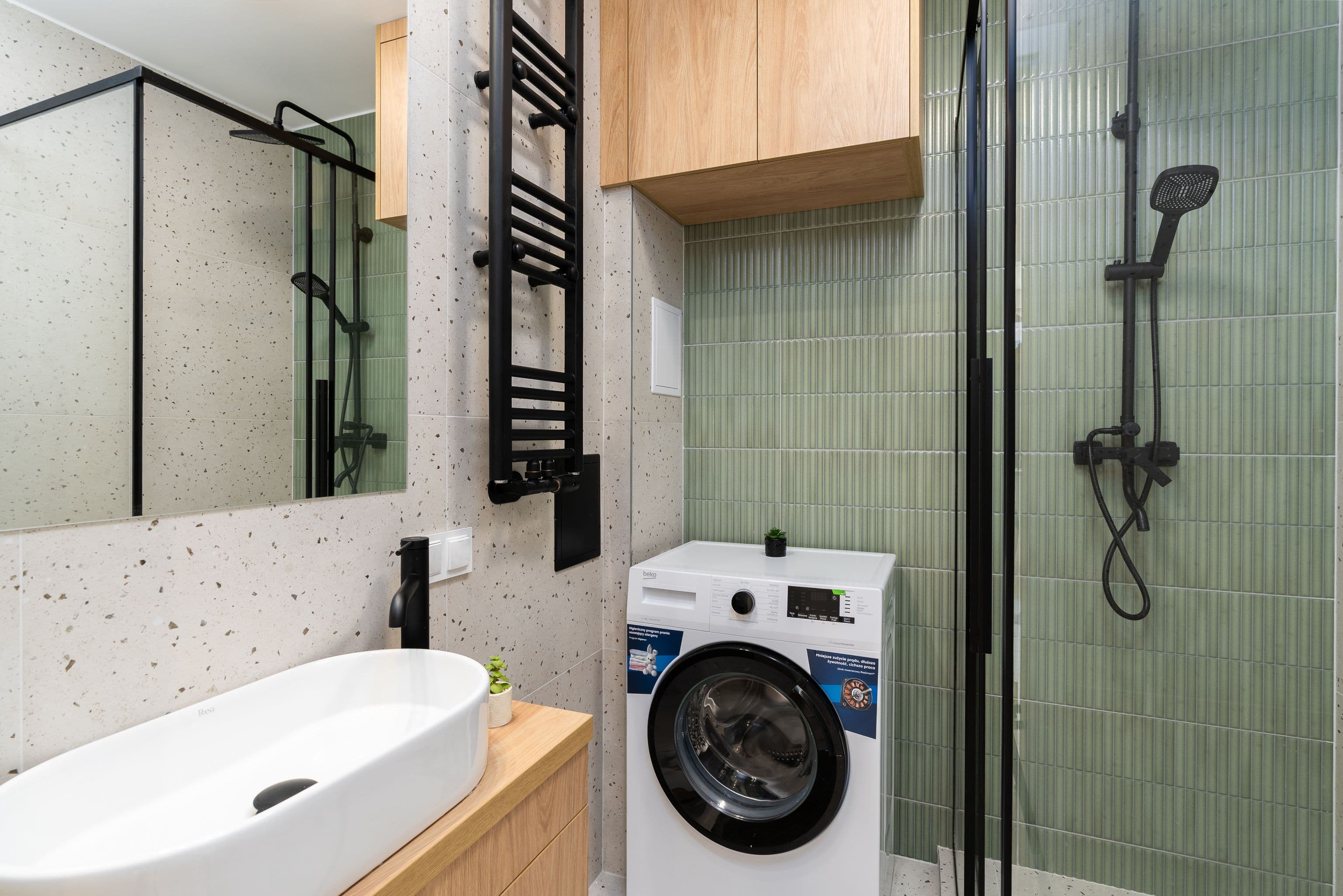 Banyo: yeşil mozaik ve terrazzo desenli fayans, siyah duş sistemi, ahşap dolap ve niş içinde çamaşır makinesi.